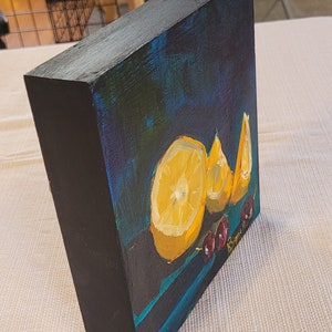 May include: A small painting on a wooden canvas with a dark blue background. The painting features three lemon slices and a few red cherries. The artist's signature is visible in the bottom right corner.