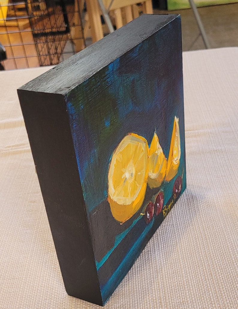 May include: A small painting on a wooden canvas with a dark blue background. The painting features three lemon slices and a few red cherries. The artist's signature is visible in the bottom right corner.