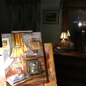 May include: A painting on an easel depicting a still life with a lamp, a book, and a framed photo. The painting is set in a room with a lamp on a table and a mirror on the wall.