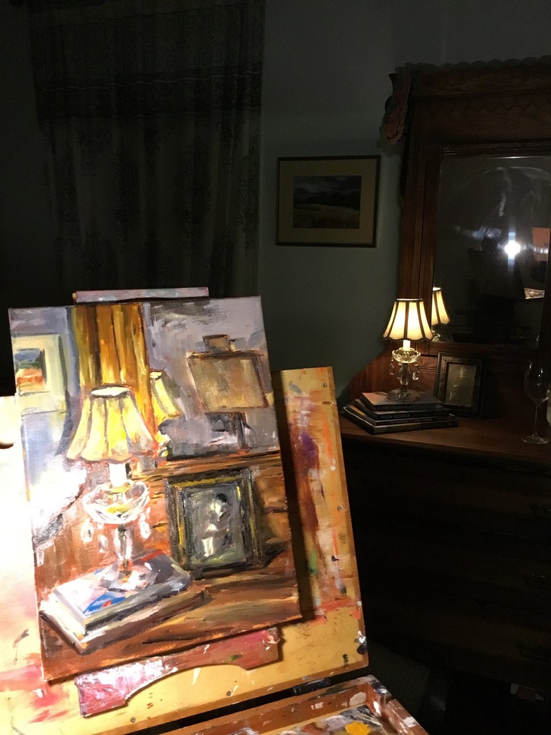 May include: A painting on an easel depicting a still life with a lamp, a book, and a framed photo. The painting is set in a room with a lamp on a table and a mirror on the wall.