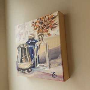 May include: A still life painting of a silver pitcher, a blue glass bottle, and a clear glass vase with orange flowers. The painting is on a canvas with a white background and a light brown table.
