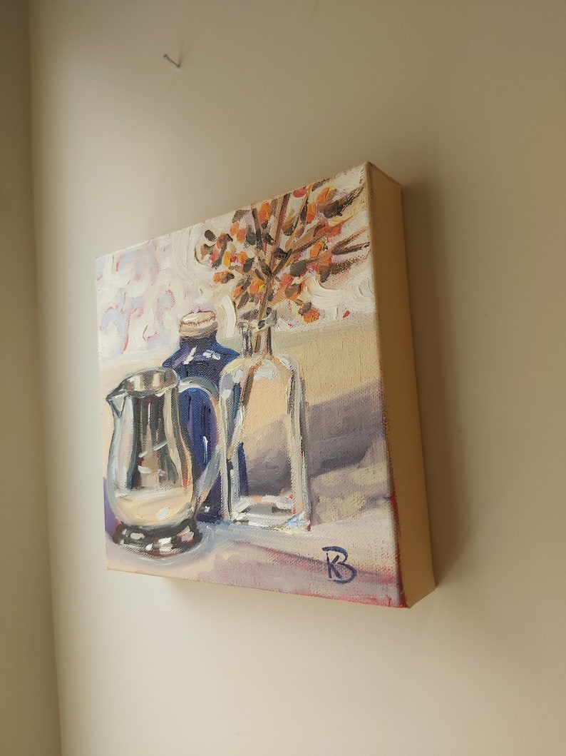 May include: A still life painting of a silver pitcher, a blue glass bottle, and a clear glass vase with orange flowers. The painting is on a canvas with a white background and a light brown table.