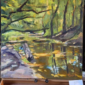 Puede incluir: Una pintura al &oacute;leo de Kathleen Bignell que representa una escena forestal serena. La obra de arte muestra una masa de agua que refleja los &aacute;rboles y el follaje. Un tronco flota en el agua. La pintura se exhibe en un caballete.