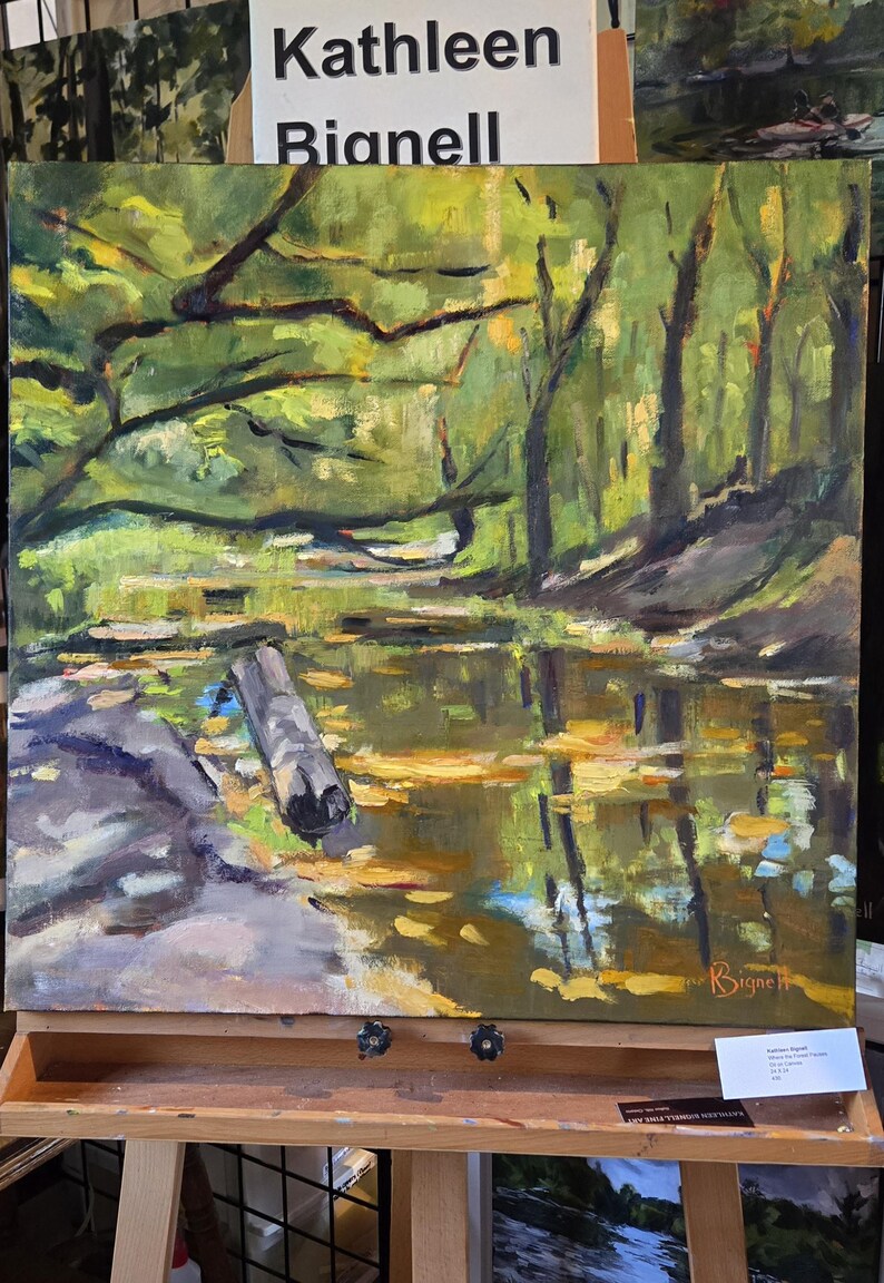 Puede incluir: Una pintura al &oacute;leo de Kathleen Bignell que representa una escena forestal serena. La obra de arte muestra una masa de agua que refleja los &aacute;rboles y el follaje. Un tronco flota en el agua. La pintura se exhibe en un caballete.