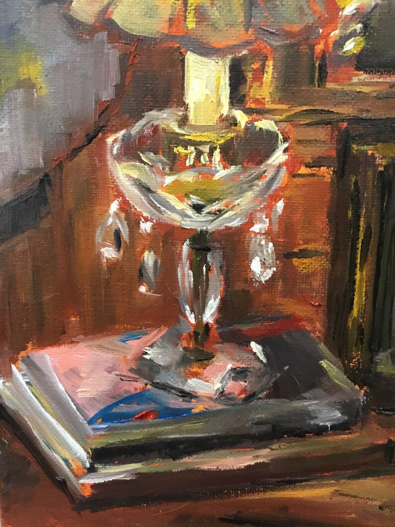 May include: A vintage-style lamp with a clear glass shade and crystal accents sits on a wooden table with a stack of books. The lamp is lit, casting a warm glow on the scene.