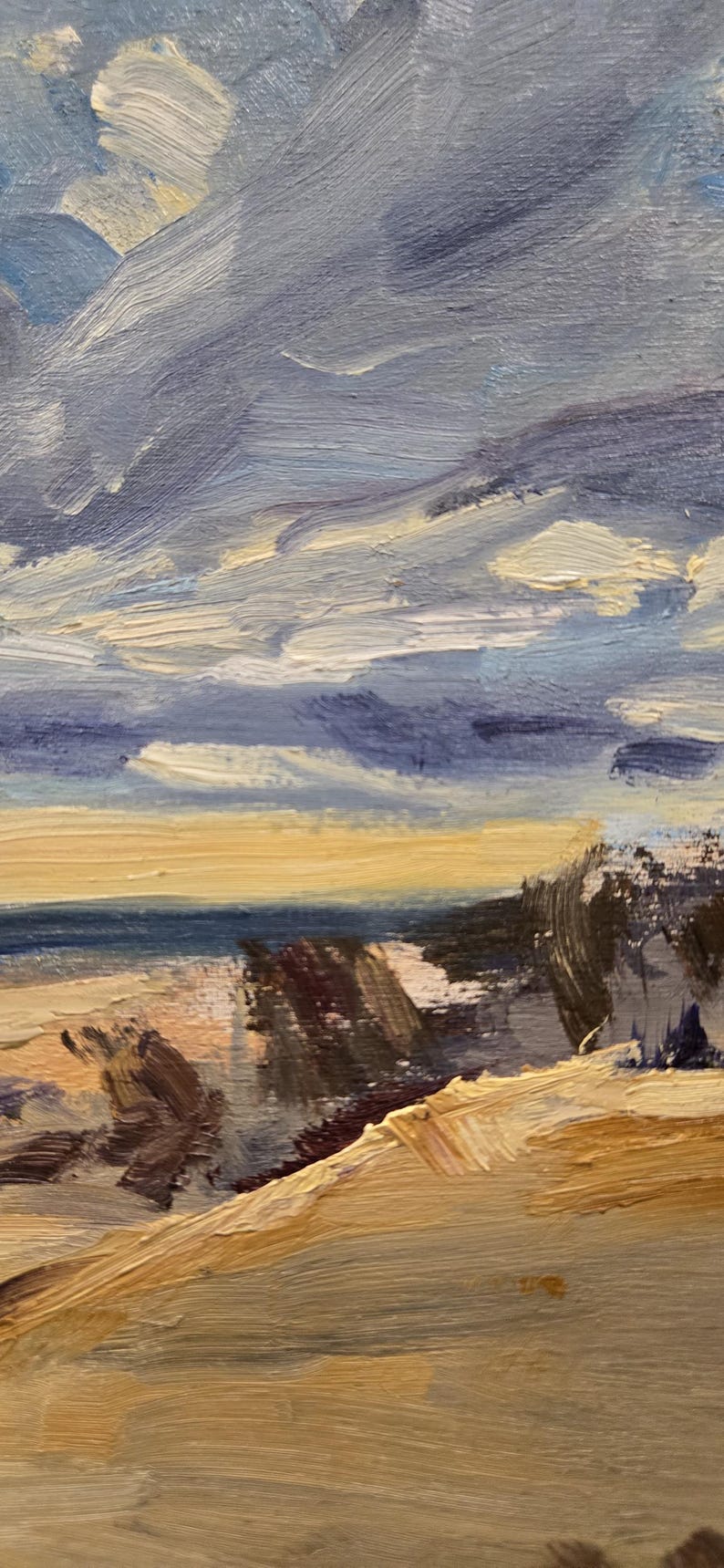 May include: An oil painting on canvas depicting a coastal landscape with thick, visible brushstrokes. The scene features a sandy foreground in shades of tan and brown, a dark blue ocean horizon, and a dramatic sky filled with swirling blue and white clouds.