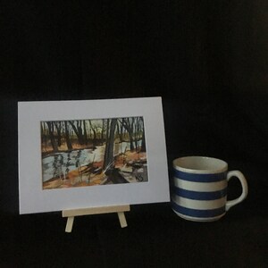 May include: A framed watercolor painting of a forest scene with a stream running through it. The painting is on a small easel and there is a blue and white striped mug in the foreground.