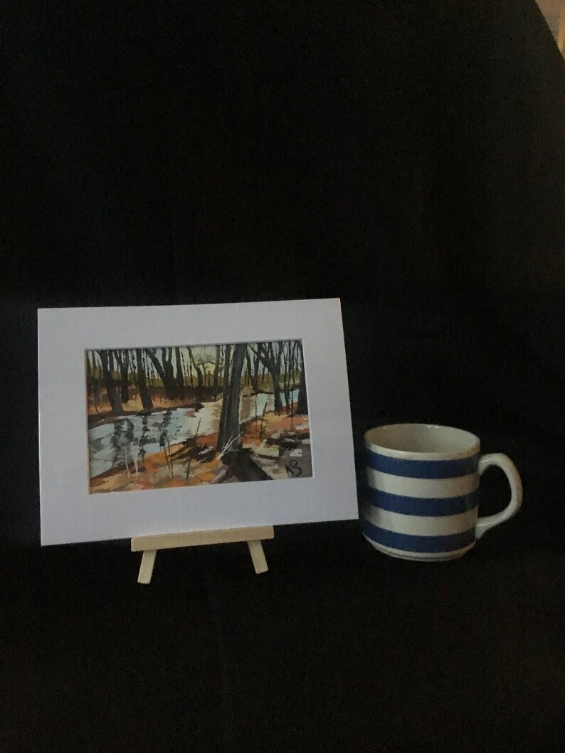 May include: A framed watercolor painting of a forest scene with a stream running through it. The painting is on a small easel and there is a blue and white striped mug in the foreground.