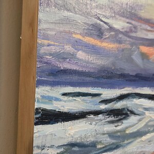 May include: Oil painting on canvas depicting a seascape with a dynamic sky. The artwork features a palette of blues, purples, and oranges, with visible brushstrokes. The painting is framed in a natural wood frame.