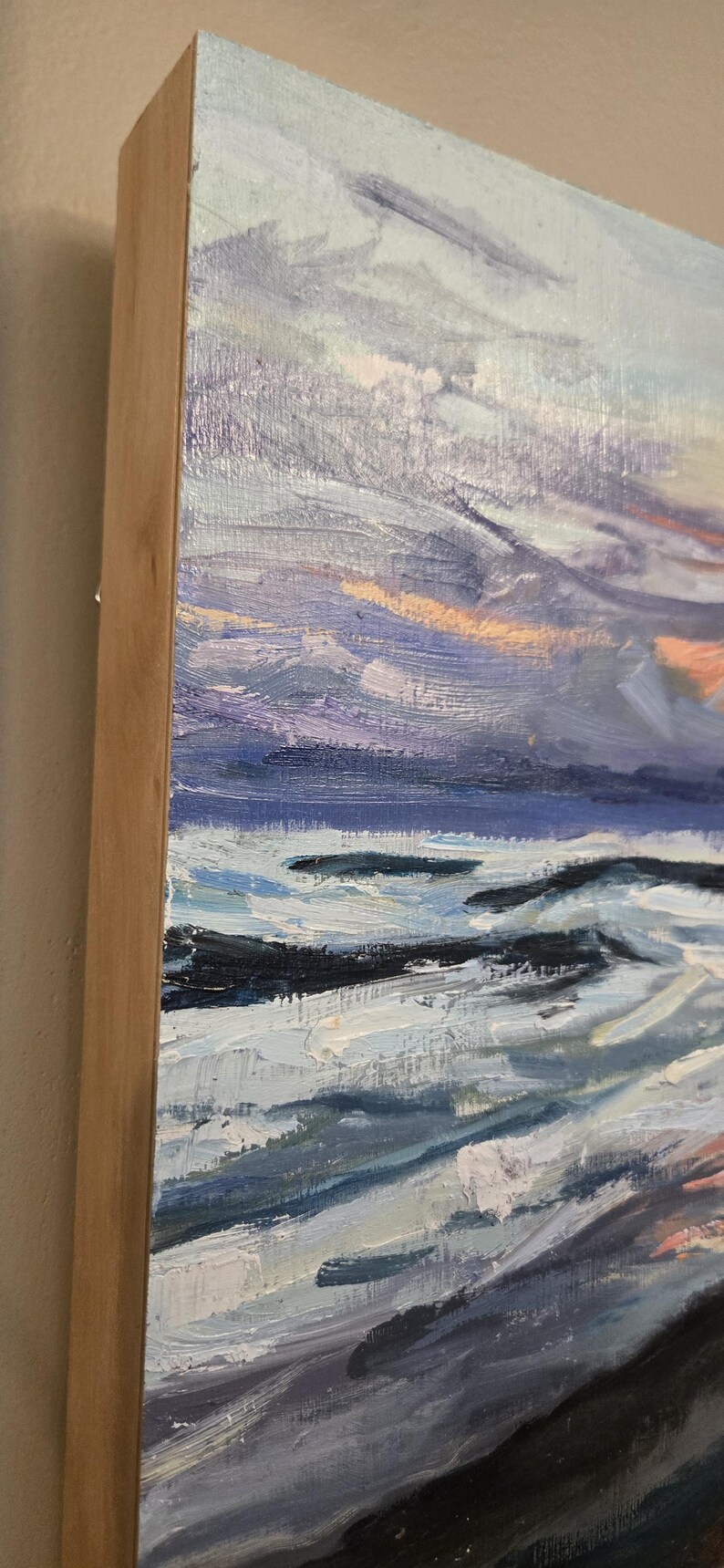May include: Oil painting on canvas depicting a seascape with a dynamic sky. The artwork features a palette of blues, purples, and oranges, with visible brushstrokes. The painting is framed in a natural wood frame.