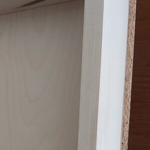 May include: Close-up of a wooden frame with a light-colored interior and a light-colored exterior edge. The frame appears to be unfinished, with visible wood grain. The side of the frame is made of a composite material.