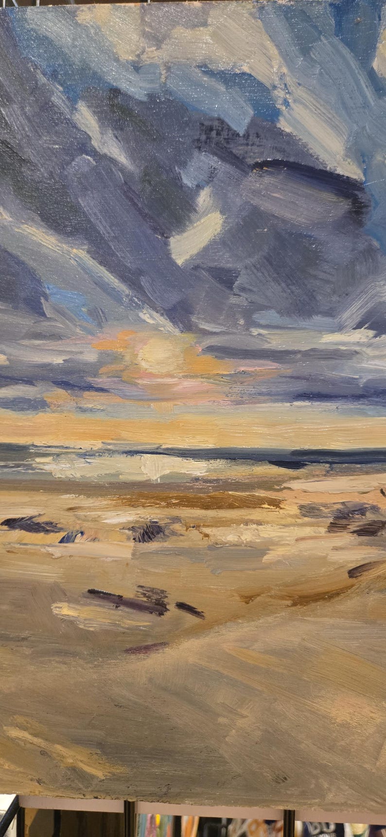 May include: An oil painting on canvas depicting a coastal landscape at sunset. The scene features a sandy beach in the foreground with visible brushstrokes in shades of beige and brown, leading to a dark blue ocean and a cloudy, textured sky.