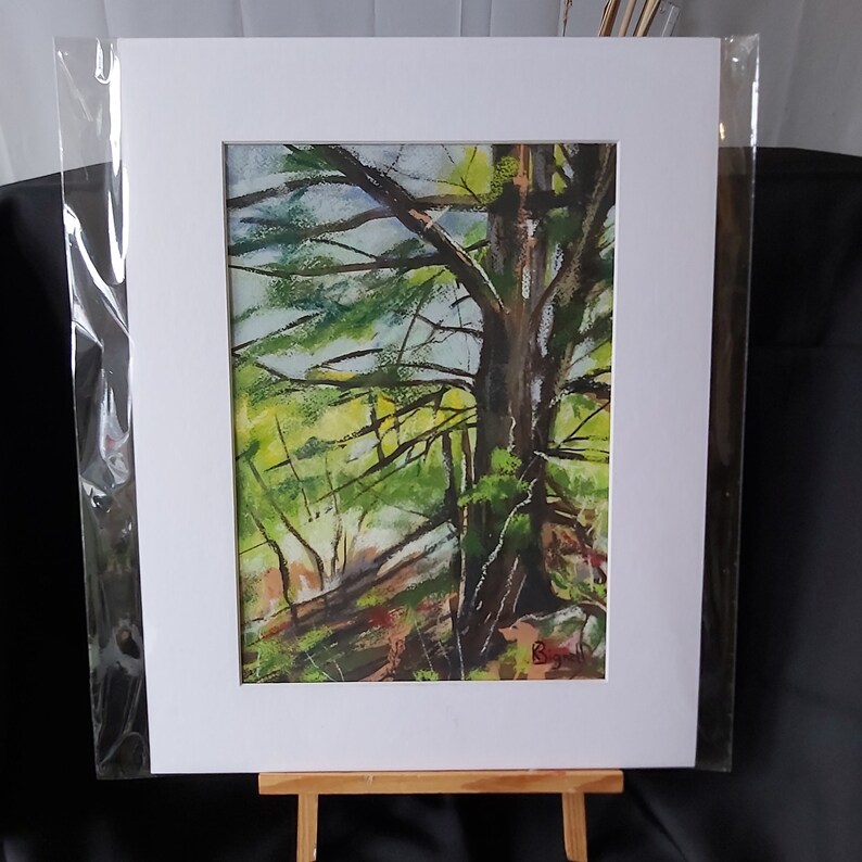 May include: A watercolor painting of a tree in a forest setting. The tree is in the foreground and the background is a blur of green and brown. The painting is framed in white and is on a wooden easel.