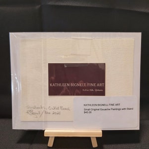 May include: A small original gouache painting of a beach scene with the title "Sandbanks - Outlet Beach" and the artist's signature "K. Bignell" and the date "Nov 2021". The painting is on a white mat and is framed in a clear plastic frame. The painting is on a small wooden easel. The painting is for sale for $45.00.