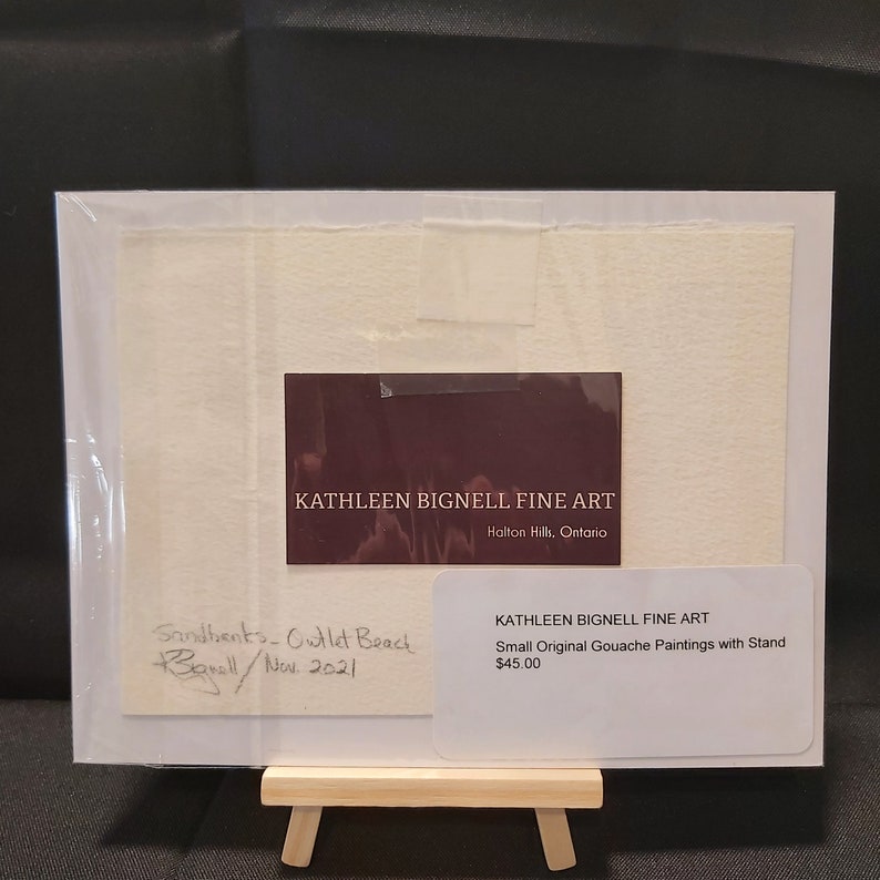 May include: A small original gouache painting of a beach scene with the title "Sandbanks - Outlet Beach" and the artist's signature "K. Bignell" and the date "Nov 2021". The painting is on a white mat and is framed in a clear plastic frame. The painting is on a small wooden easel. The painting is for sale for $45.00.