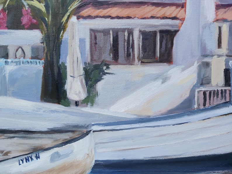 May include: Two white wooden boats with a blue stripe on the bottom. The boat in the foreground has the text "LYNX W" on the side. The boats are docked in front of a white building with a red tile roof.