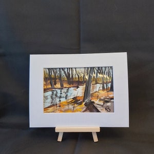 May include: A framed painting of a forest scene with a stream running through it. The trees are bare and the water is a light blue. The painting is in a white frame and is on a small wooden easel.