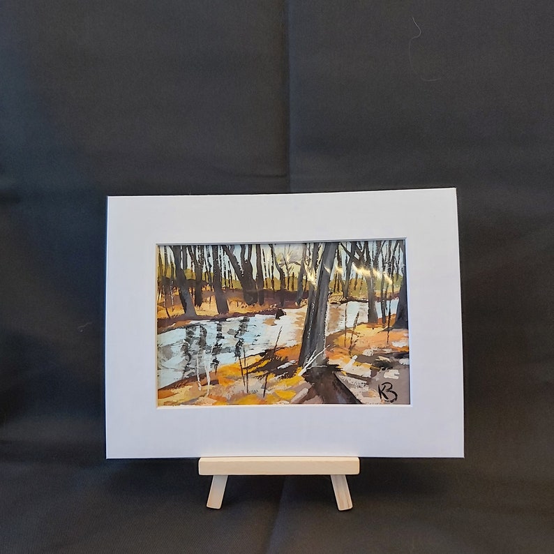 May include: A framed painting of a forest scene with a stream running through it. The trees are bare and the water is a light blue. The painting is in a white frame and is on a small wooden easel.