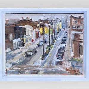 May include: An oil painting of a city street scene, framed in white. The artwork depicts a road with cars, streetlights, and buildings in shades of brown, gray, and white. The artist's signature is visible in the lower right corner.