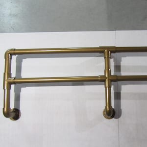 May include: A golden-colored, industrial-style handrail made of connected pipes and fittings. The handrail has a horizontal bar design with two parallel bars and vertical supports. The metal has a shiny finish and is mounted on a white surface.