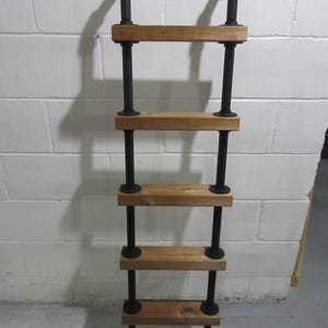 May include: A rustic, industrial-style ladder shelf. The shelf features four wooden shelves supported by black metal pipes and fittings. The ladder is designed to be mounted on a wall, with two black metal brackets at the top. The shelves are made of light-colored wood.