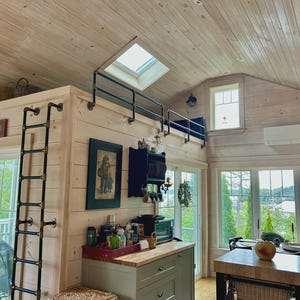 May include: Interior shot of a light-filled kitchen and living space with wood paneling and a loft area. A black metal ladder leads to the loft, and a small railing is installed above the kitchen. The room features large windows and a dining table.