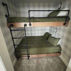 May include: A bunk bed with a wooden frame and black metal pipe accents. The beds have olive green bedding and pillows. A ladder made of black metal pipes provides access to the top bunk. The room has plaid wallpaper.