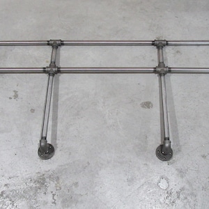 May include: Industrial metal pipe shelf with a rectangular design. The shelf has horizontal bars connected by elbow joints and vertical supports ending in circular flanges. The metal has a silver finish.