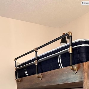 May include: A loft bed with a dark blue mattress and a black metal railing. The railing is constructed with black pipes and brass fittings. A small black lamp is mounted on the side.