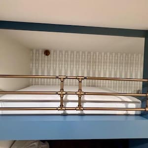 May include: A bunk bed featuring a brass pipe safety rail. The bed frame is painted blue, and the mattress is white. The wall behind the bed has patterned wallpaper. A small, round, brass-coloured object is mounted on the wall.