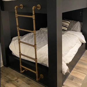 May include: A black bunk bed frame featuring a metallic gold industrial pipe ladder attached to the side. The bed includes white bedding with a textured quilt and a striped pillow. The structure is built with dark, solid vertical posts.