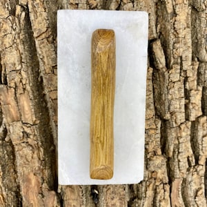 May include: A wooden pestle with a light brown finish rests on a white rectangular mortar. The pestle is cylindrical with a rounded end. The background is a textured brown surface.