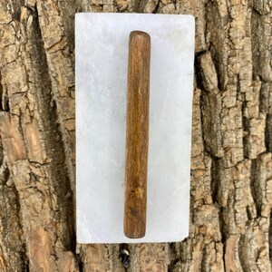 May include: A wooden rolling pin rests on a white rectangular marble slab. The rolling pin is a medium brown color and has a smooth, rounded shape. The marble slab has a polished surface.