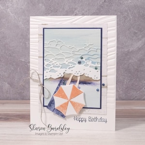May include: A handmade greeting card with a beach theme. The card features a paper-cut wave design, a small orange and white umbrella, and the words "Happy Birthday." The card is white with a textured border and a blue and white background.
