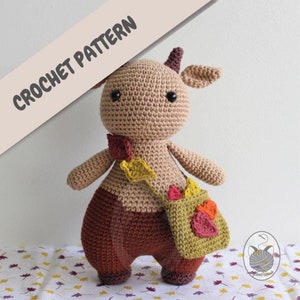 May include: Crochet pattern for a brown and tan amigurumi deer with a small green bag and fall leaves. The deer has black eyes and a brown nose.
