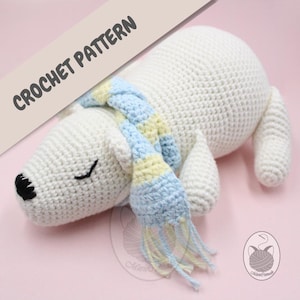 Puede incluir: Oso polar de crochet blanco con una bufanda a rayas azules y amarillas con borlas. El oso está acostado con los ojos cerrados. Patrón de crochet.