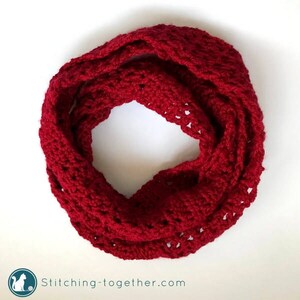 May include: A red crocheted infinity scarf with a lacy pattern. The scarf is folded in half and laid flat on a white surface.