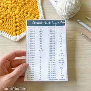 May include: A crochet hook size chart with metric and US sizes listed in columns. The chart is printed on a white card with a purple border. The chart is being held by a hand.
