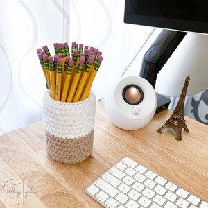 May include: A handmade crochet pencil holder in white and tan, filled with yellow pencils with pink erasers. The pencil holder sits on a wooden desk next to a white speaker, a miniature Eiffel Tower, and a keyboard.