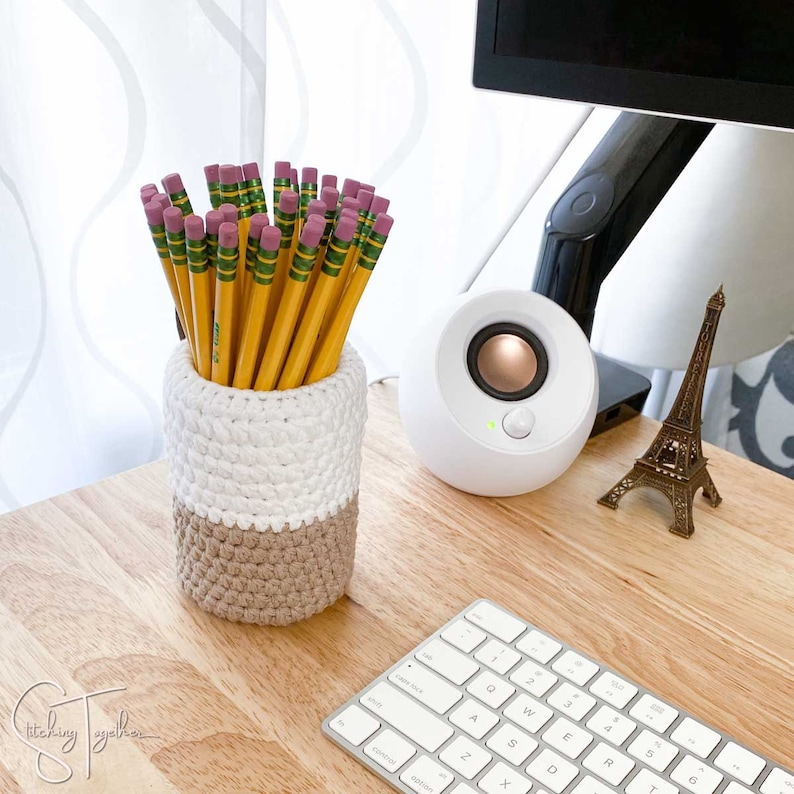 May include: A handmade crochet pencil holder in white and tan, filled with yellow pencils with pink erasers. The pencil holder sits on a wooden desk next to a white speaker, a miniature Eiffel Tower, and a keyboard.