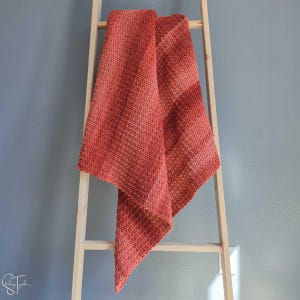 May include: A coral-coloured knitted blanket draped over a wooden ladder. The blanket has a textured stitch pattern and a gradient effect, changing from a darker shade to a lighter hue. The blanket is a cosy home decor item.