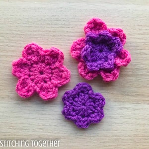 May include: Three crocheted flowers, one pink and two purple, on a woodgrain surface. The pink flower is the largest, and the two purple flowers are smaller.