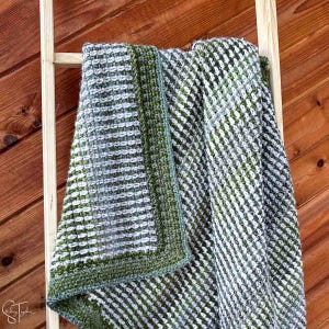 May include: A green and white crocheted throw blanket with a textured pattern. The blanket is folded and draped over a wooden ladder.