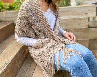 May include: A woman wearing a beige crochet shawl with fringe. The shawl has a textured pattern and is worn over a white shirt and blue jeans.