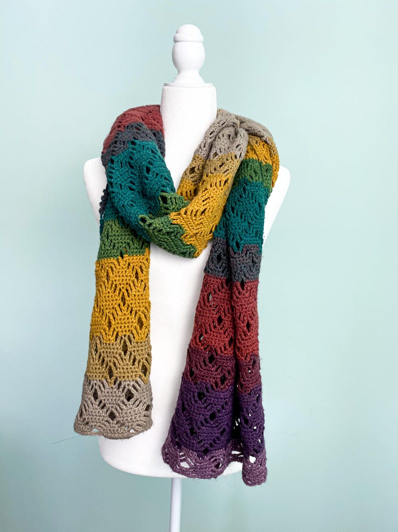 Puede incluir: Una bufanda de ganchillo multicolor con un patr&oacute;n de encaje. La bufanda est&aacute; hecha en tonos de morado, rojo, naranja, amarillo, verde y gris.