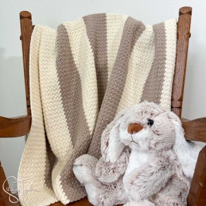 Könnte beinhalten: Eine gestrickte gestreifte Decke in Beige und Weiß liegt über einem Holz-Schaukelstuhl. Ein Plüsch-Hase in Braun und Weiß sitzt auf der Decke.