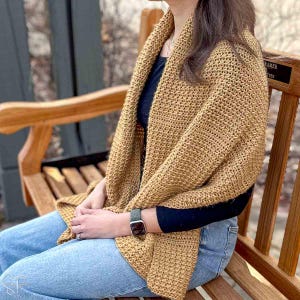 Puede incluir: Un chal de ganchillo color beige, drapeado sobre los hombros de una persona. El chal tiene un patrón de puntada texturizada y se usa sobre una parte superior oscura y jeans azules. La persona está sentada en un banco de madera.