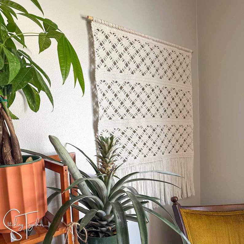 May include: Cream-colored macrame wall hanging with a geometric diamond pattern and fringe. The wall decor is mounted on a wooden dowel. The macrame is displayed on a white wall, with a pineapple plant and other greenery in the foreground.
