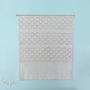 May include: Cream-colored macrame wall hanging with a geometric pattern and fringe. The wall decor is mounted on a wooden dowel. The background is a light blue wall. The text "Stitching Together" is in the bottom left corner.