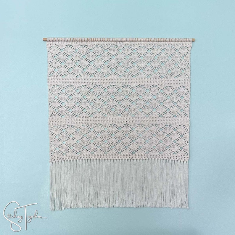 May include: Cream-colored macrame wall hanging with a geometric pattern and fringe. The wall decor is mounted on a wooden dowel. The background is a light blue wall. The text "Stitching Together" is in the bottom left corner.